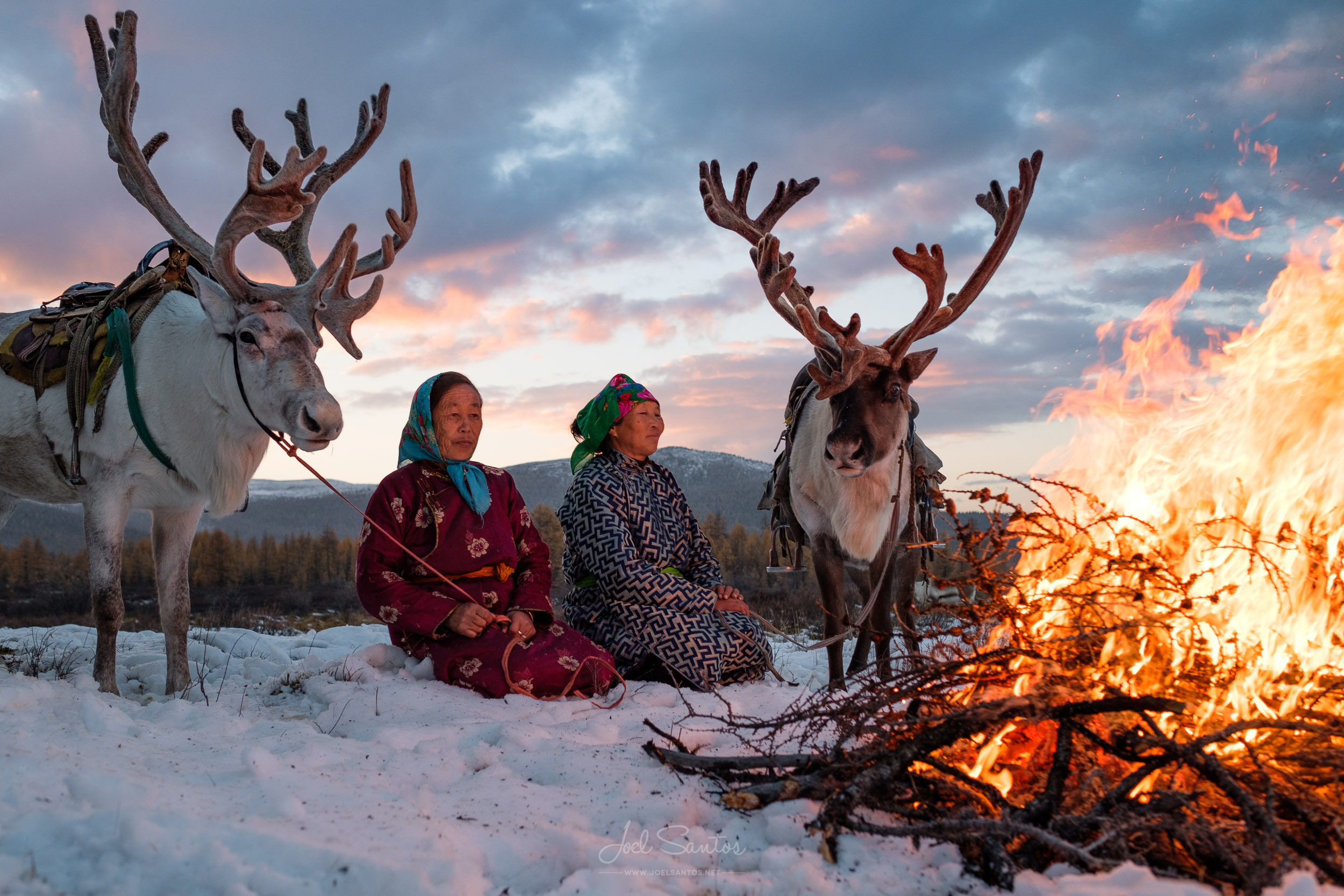 Two Hanai herders sit around a campfire in the snow with their two reindeers