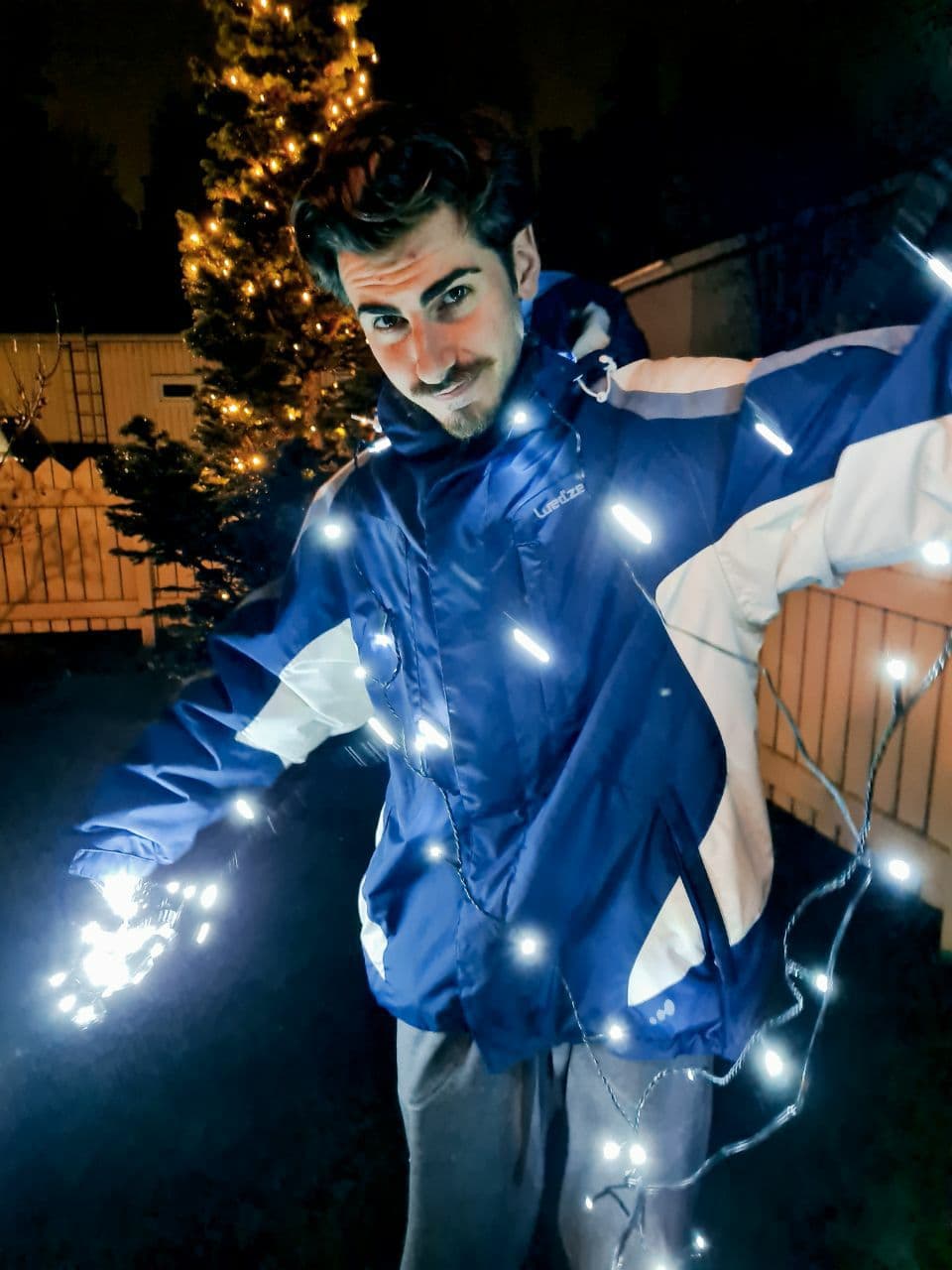 Clockwork in a blue winter jacket holding Christmas lights in a garden by night